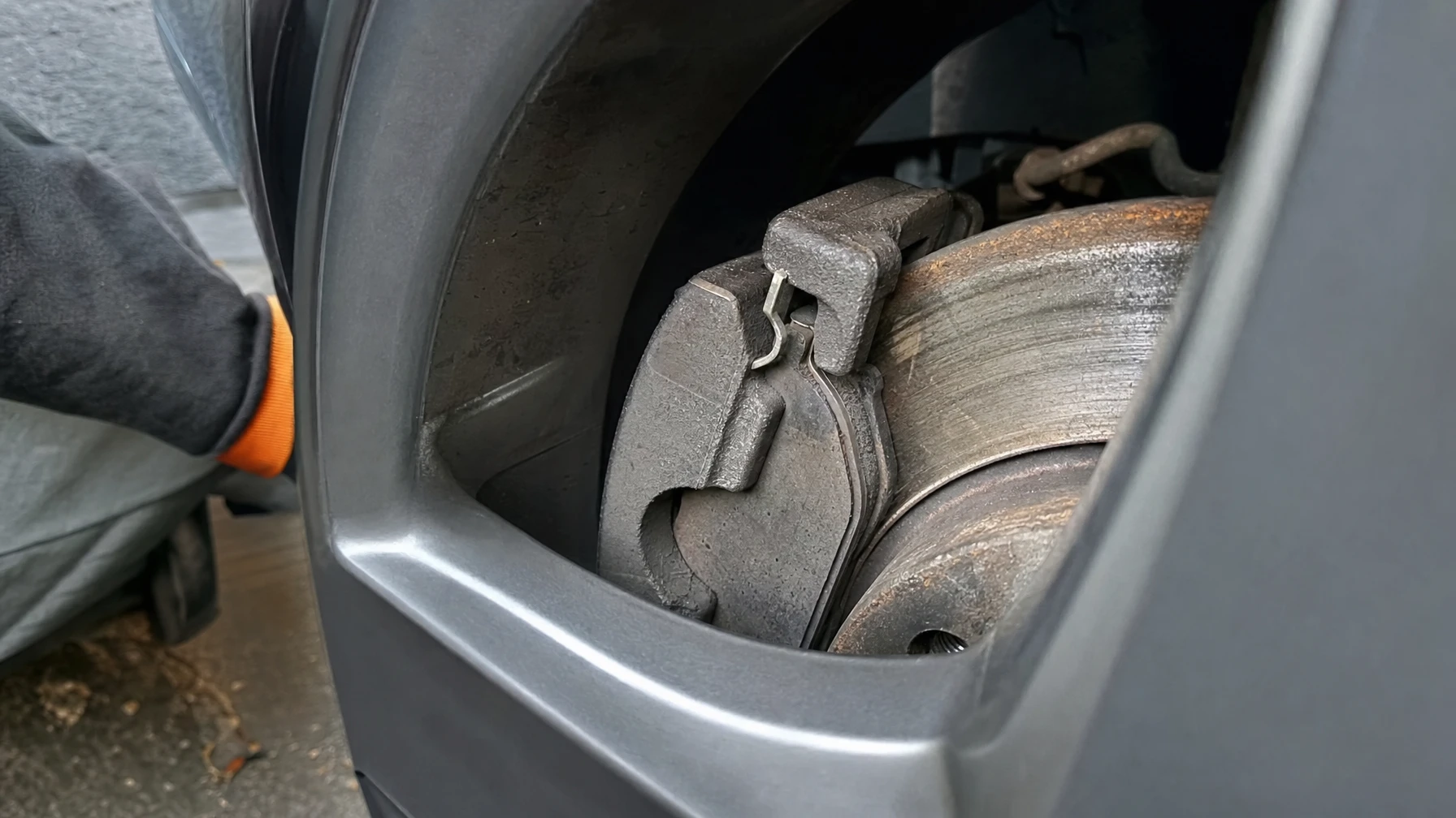 A worn brake pad and caliper viewed through a car's wheel spokes, pressed against the rotor