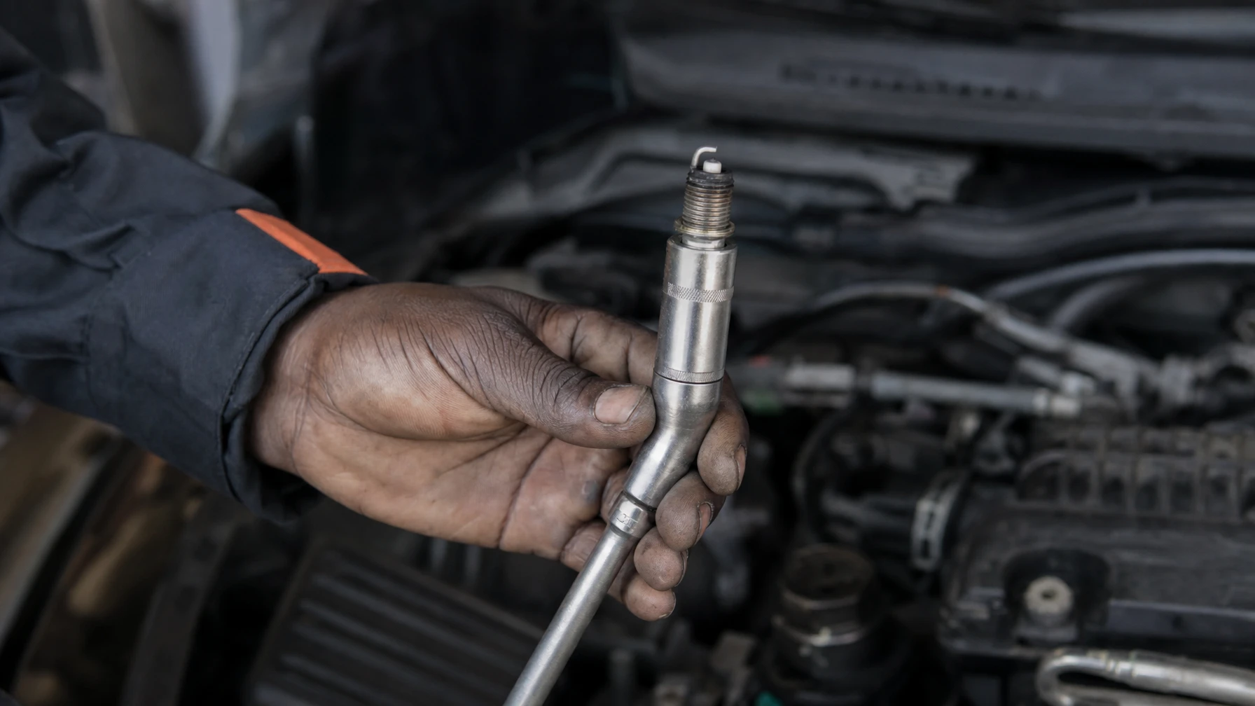 A mechanic holding a spark plug on a socket extension next to a car's engine
