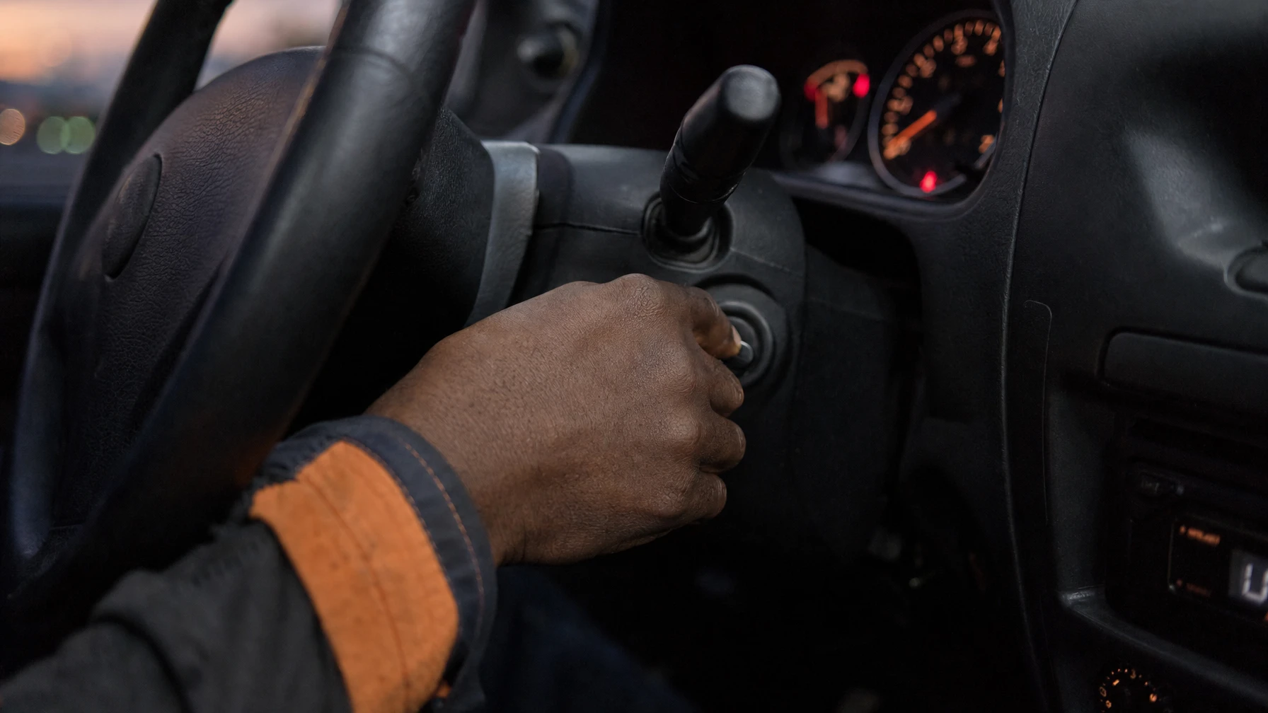 A hand turning a car key in the ignition at dusk, dashboard gauges glowing